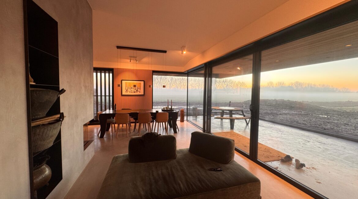 Moderne woonkamer met grote glazen schuifdeuren die uitzicht bieden op een open landschap bij zonsopkomst, een minimalistische eettafel met houten stoelen en een comfortabele groene loungebank