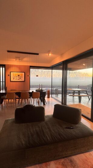 Moderne woonkamer met grote glazen schuifdeuren die uitzicht bieden op een open landschap bij zonsopkomst, een minimalistische eettafel met houten stoelen en een comfortabele groene loungebank