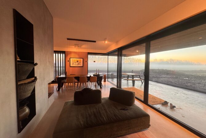Moderne woonkamer met grote glazen schuifdeuren die uitzicht bieden op een open landschap bij zonsopkomst, een minimalistische eettafel met houten stoelen en een comfortabele groene loungebank