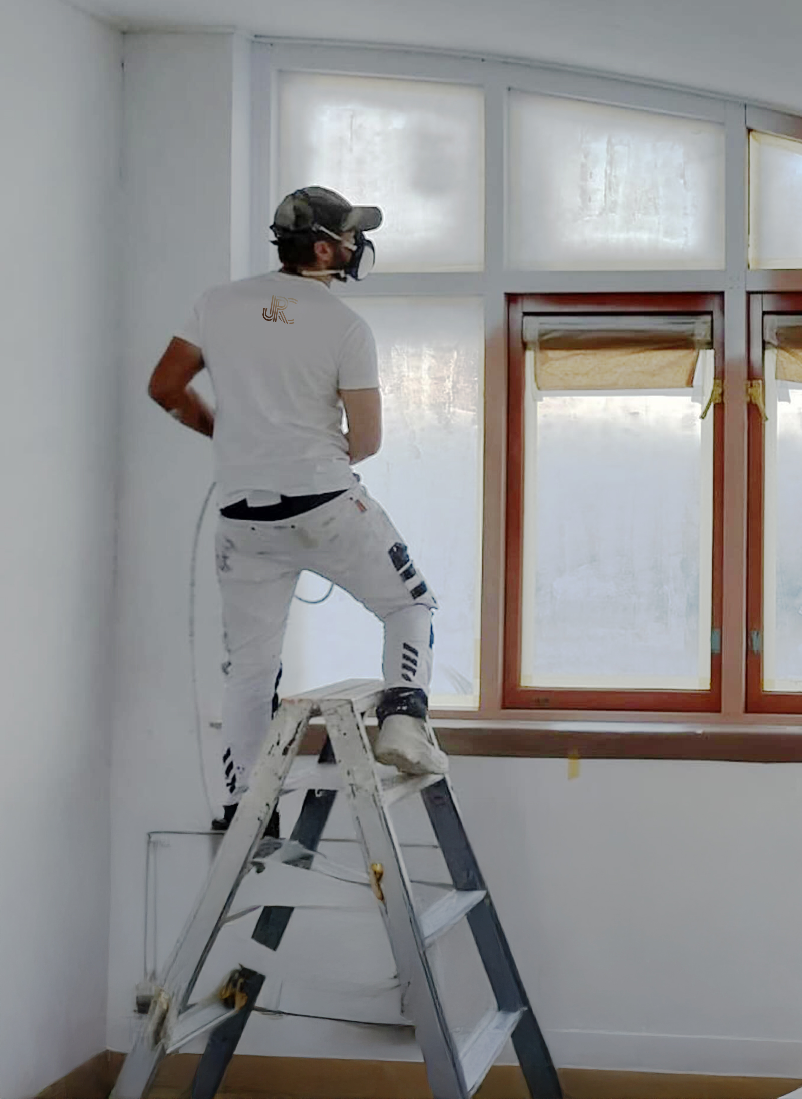 Vakspecialist van JRC Afbouw spuit verf op een muur in een lichte ruimte, gekleed in een wit uniform voorzien van het JRC Afbouw-logo, werkend bij een groot raam