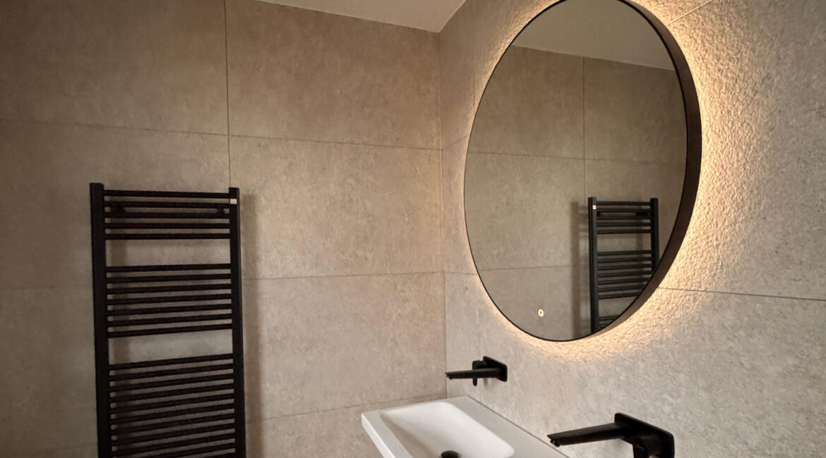 Tijdloos en strak badkamerontwerp. Moderne badkamer met strakke tegels en een ronde spiegel, afgewerkt door JRC Afbouw.