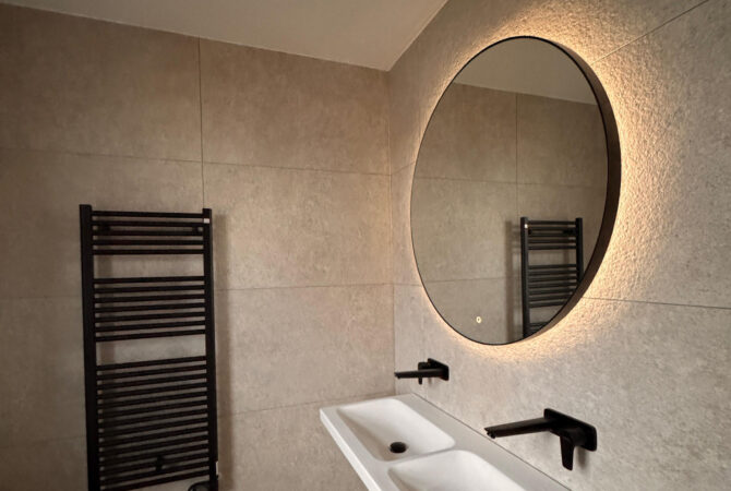Moderne badkamer met strakke tegels en een ronde spiegel, afgewerkt door JRC Afbouw.